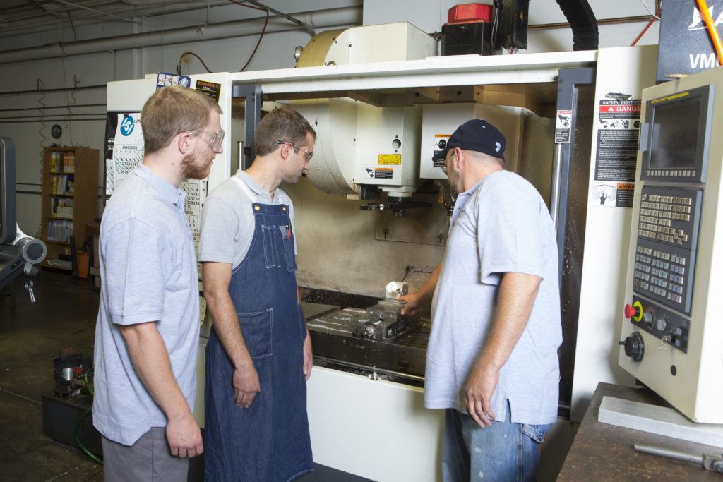 Three technicians observing and operating a CNC machining center, showcasing advanced manufacturing processes at Monsees Group, emphasizing efficiency and manufacturability in precision job shop operations.