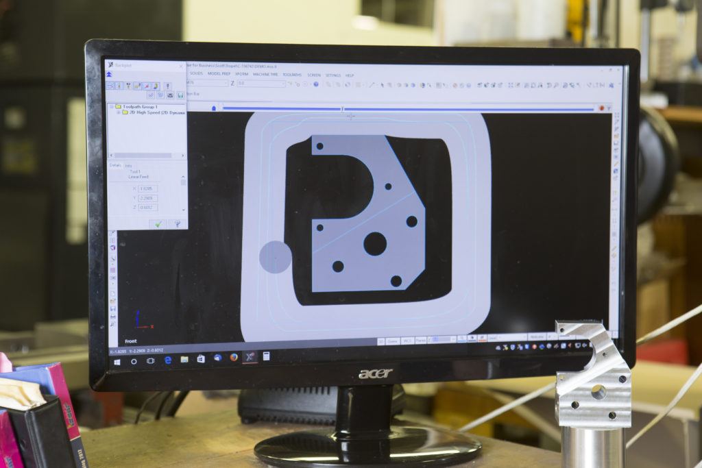 Computer monitor displaying Mastercam software interface with CNC machining design, featuring a part outline with various holes and contours, alongside a machined metal component on the workbench in a manufacturing environment.