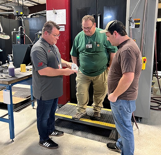Three men discussing machining processes in a workshop, one holding a component, surrounded by CNC machines and tools, illustrating collaboration and training related to Mastercam software at Avans Machine Inc.