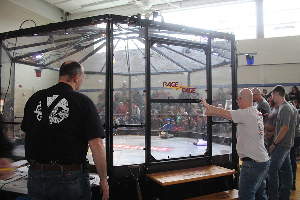 BattleBot competition arena with spectators, featuring a clear cage labeled "RAGE CAGE," as participants prepare for a robot battle in a school gymnasium setting.