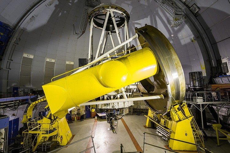 Large yellow telescope inside observatory, showcasing precision engineering for astronomical research, with intricate structural components and machining setups.