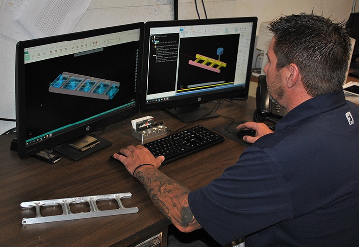CNC supervisor programming winder frame part using Mastercam CAD/CAM software on dual monitors, with a machined part in the foreground, illustrating advanced manufacturing processes at Stainless Fittings Group (SFG).