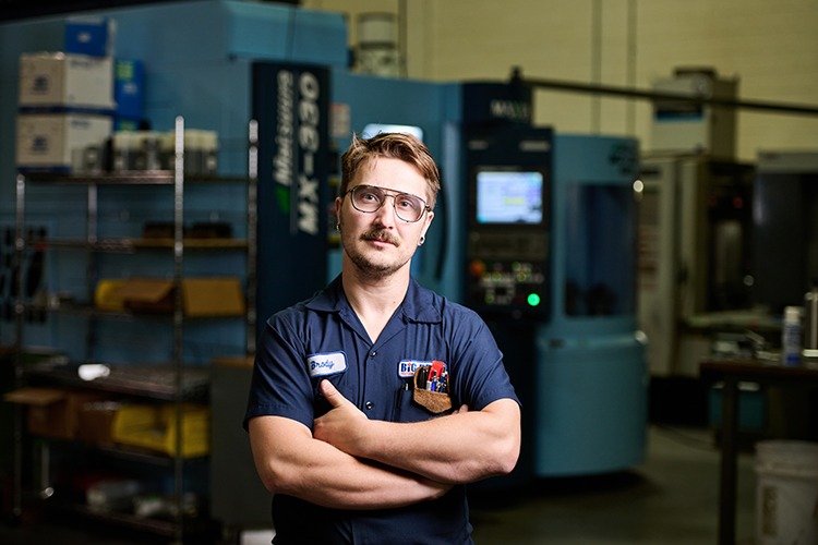 Brody Huk, Manufacturing Engineer at Big Drum Precision Machining, standing confidently in front of advanced multiaxis machining equipment in a precision machining facility.