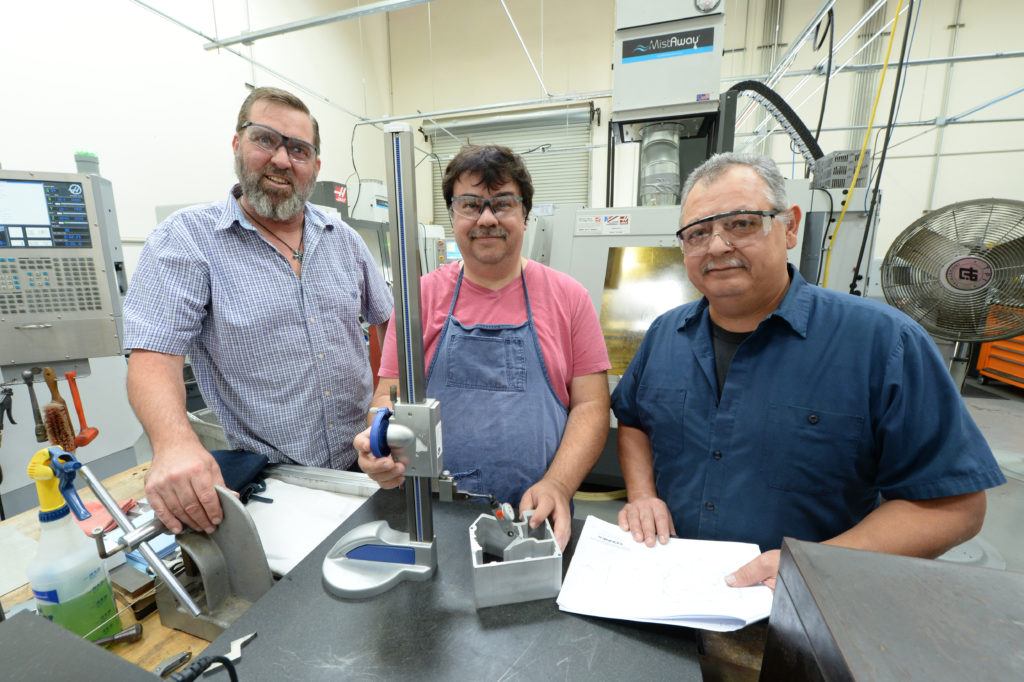 Three machinists in a workshop, wearing safety glasses and working with tools and a prototype part, emphasizing collaboration and precision in engineering for medical device manufacturing.
