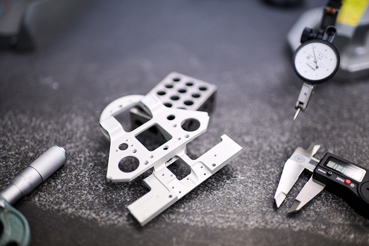Precision machined metal part with multiple holes and contours, accompanied by calipers and a dial gauge, on a workbench, illustrating high-precision manufacturing for medical automation components.