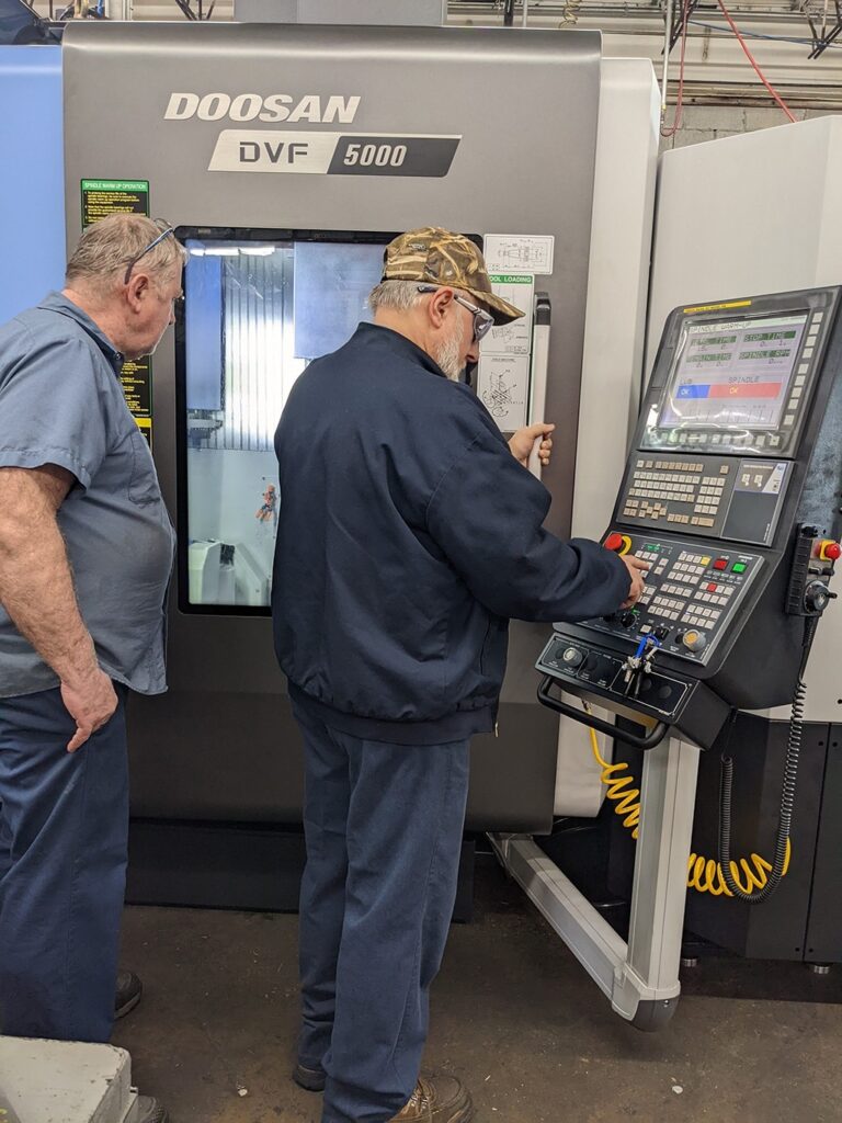 Two machinists programming a part on a Doosan DVF 5000 machine in a manufacturing facility, showcasing the integration of Mastercam CAD/CAM software for enhanced production efficiency.