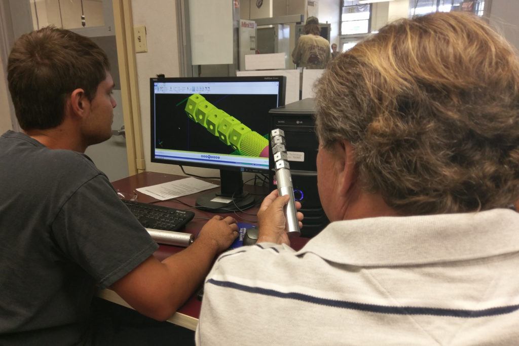 Students engaged in CAD/CAM programming using Mastercam software, analyzing a 3D model on a computer screen, with one student holding a measuring tool.
