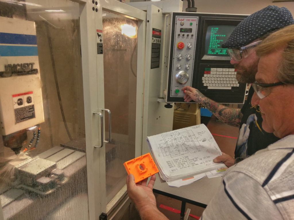 Two individuals examining a blueprint while operating a CNC milling machine, with one holding an orange plastic part, showcasing hands-on learning in the Machine Technology Program at San Diego City College.