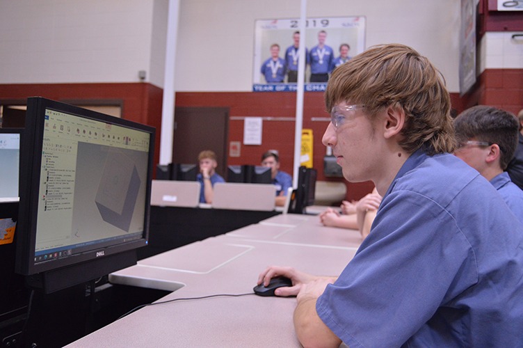 Precision machining student using Mastercam software to program toolpaths, focused on computer screen, with classmates in background.