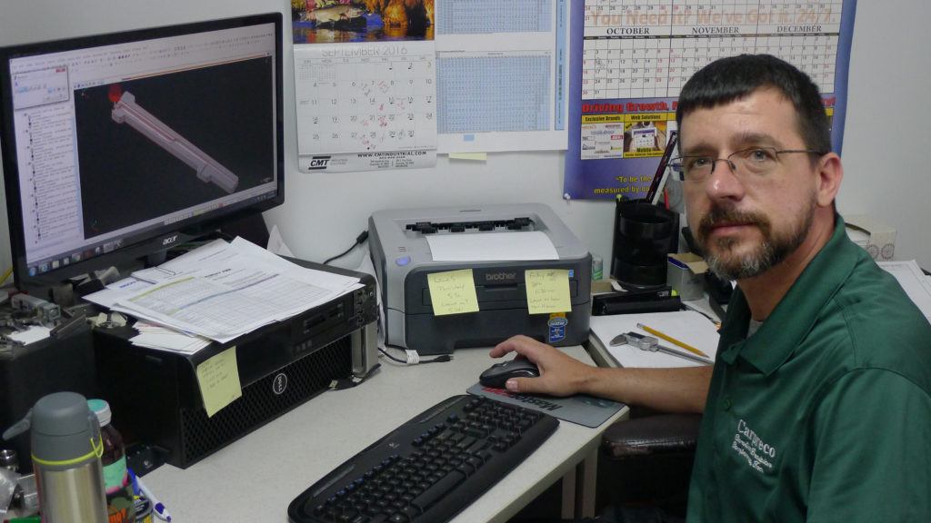 Process engineer at workstation using Mastercam software for CNC programming, with a 3D model displayed on the monitor, surrounded by office supplies and a calendar.