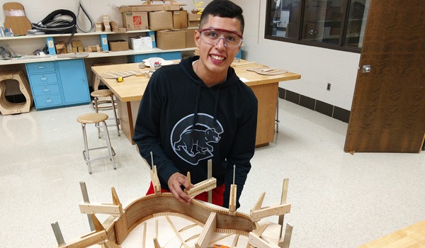 Student wearing safety glasses working on guitar assembly in woodworking classroom, surrounded by tools and materials for guitar-making project.